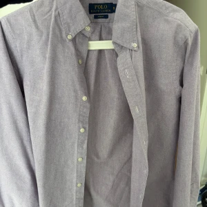 Ljuslila skjorta från Polo Ralph Lauren - Säljer en stilren ljuslila skjorta från Polo Ralph Lauren i slim fit-modell. Skjortan har klassisk button down-krage, broderad logga på bröstet och knäppning framtill. Perfekt för dig som gillar en clean och tidlös look.