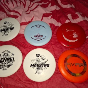 Samling av discgolf-discar - Sex olika discgolf-discar: två vita Maestro från Discmania, en vit Sensei från Discmania, en ljusblå Hope från Latitude 64, en röd Caiman från Innova och en orange Aviar från Innova. Discarna har olika tryck och färger, perfekta för discgolfspelare.