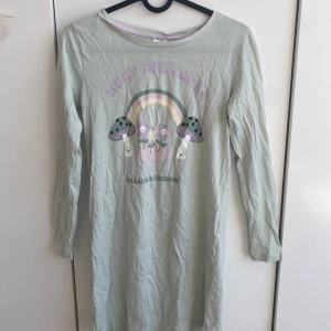 Ljusgrön långärmad klänning med tryck från Kappahl - Ljusgrön långärmad nattlinne från Kappahl med gulligt tryck av regnbåge, svampar och texten 'Keep Dreaming'. Mjuk bomullskänsla och avslappnad passform, perfekt för en skön pyjamas.