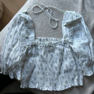 Blommig croppad blus från H&M - Säljer en söt blommig croppad blus från H&M i vitt med ljusgröna och lila blommor. Blusen har långa puffärmar, smockad midja och knytband i nacken. Perfekt för vår och sommar! Sparsamt använd 🌸🌸