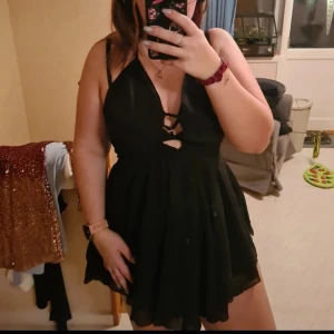 Svart kort klänning med v-ringning och smala axelband - Säljer en svart kort klänning med v-ringning och smala axelband. Klänningen har omlottliknande överdel med dekorativa banddetaljer framtill och en lätt utställd kjol. Perfekt för dig som gillar en stilren och enkel look. Aldrig använd, nypris 159kr