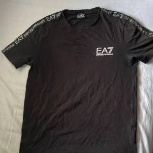 Svart t-shirt från Emporio Armani EA7 - Säljer en svart t-shirt från Emporio Armani EA7 med logga på bröstet och tryckt text längs axlarna. Klassisk rund hals och korta ärmar. Perfekt för en stilren och sportig look.