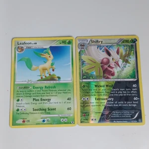 Pokémonkort Leafeon och Shiftry - Två Pokémonkort: Leafeon (grön, med gul ram) och Shiftry (grön, med gul ram). Båda korten har illustrationer av respektive Pokémon och innehåller attacker och energisymboler. Perfekt för samlare eller Pokémon-entusiaster.