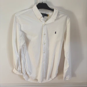 Vit skjorta från Polo Ralph Lauren - Klassisk vit skjorta från Polo Ralph Lauren med broderad logga på bröstet. Skjortan har lång ärm, knappar framtill och krage. Perfekt för en stilren look. Det är en deodorant fläck i armhålan men inget som syns om man inte vet 