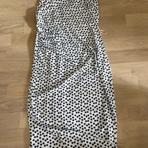 Vit prickig långklänning med smala axelband - Säljer en vit långklänning med svarta prickar och smala axelband. Klänningen har en tight passform och är tillverkad i ett mjukt, lätt material som känns svalt mot huden. Perfekt för sommarens varma dagar eller studentfirande.