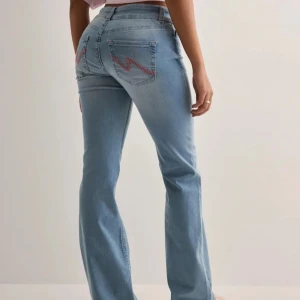 Nelly low waist jeans - Säljer ett par ljusblå jeans med bootcut passform och klassisk femficksdesign. Jeansen har en snygg broderad detalj i rosa på bakfickan och stängs med knapp och dragkedja. Perfekta för en avslappnad och trendig look. OBS: lite slitna längst ner på benen! Nypris 699kr🩷