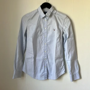 Ljusblå skjorta från GANT - Säljer en klassisk ljusblå skjorta från GANT med broderad logga på bröstet. Skjortan har lång ärm, knappar framtill och krage. Perfekt för en stilren look och passar till många tillfällen.