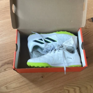 Vita Adidas Copa fotbollsskor med neongul sula - Säljer ett par vita Adidas Copa fotbollsskor med neongul sula och svarta ränder på sidan. Skorna har snörning och är perfekta för spel på konstgräs. Inte original kartong