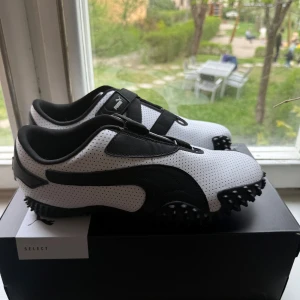 Puma Mostro Perf vita och svarta sneakers - Säljer ett par Puma Mostro Perf sneakers i vitt och svart med perforerat syntetmaterial och svarta detaljer. Skorna har kardborreband och en unik sula med gummidobbar för extra grepp. Perfekta för dig som gillar sportig och modern stil.
