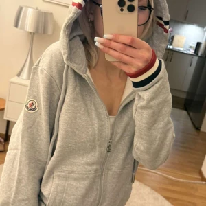 Grå hoodie med dragkedja från Moncler - Snygg grå hoodie med dragkedja från Moncler. Jackan har huva med röd, vit och blå randdetalj samt matchande muddar. Klassisk Moncler-logga på ärmen och två fickor framtill. Perfekt för vilken stil som helst! Inte använd endast tagit bilder i tröjan. 