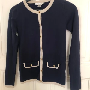 Mörkblå kofta med beige detaljer från Mango - Säljer en mörkblå kofta från Mango med beige kantband och guldknappar. Koftan har två små fickor framtill med dekorativa knappar och långa ärmar. Perfekt för lager-på-lager och stilren look.