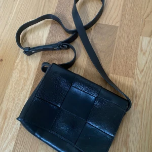 Svart axelväska i skinn med flätad design från Lindex - Säljer en svart axelväska i äkta skinn med flätad framsida och justerbart axelband. Perfekt storlek (20x15 cm) för det viktigaste och enkel att matcha till olika outfits.