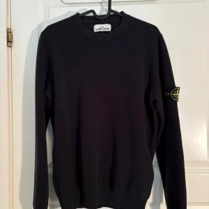 Svart stickad tröja från Stone Island - Säljer en svart stickad tröja från Stone Island med klassisk rund hals och ribbade muddar. Tröjan har den ikoniska loggan på vänster ärm och är perfekt för dig som gillar stilrena plagg med streetkänsla.