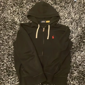 Svart hoodie från Polo Ralph Lauren - Snygg svart hoodie från Polo Ralph Lauren med dragkedja och huva. Den har en röd broderad logga på bröstet och vita snören vid huvan. Perfekt för en avslappnad stil. Köpt från Zalando för 2200 säljer för 1200 