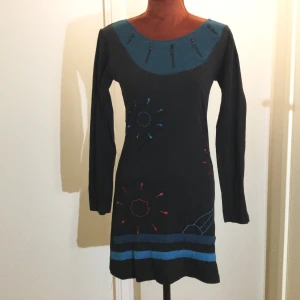  klänning med broderade detaljer S - Unik klänning med lång ärm och rund halsringning. Klänningen har broderade detaljer i blått och rött samt dekorativa dragkedjor vid halsen. Två blå ränder nertill ger extra stil. Tillverkad i mjukt material, perfekt för dig som vill sticka ut. Klänning Svart med mönster Strl: S