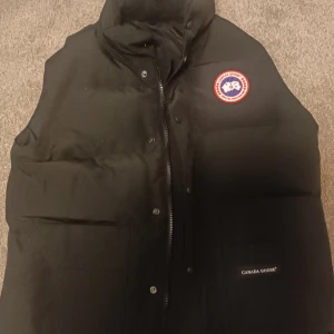 Svart dunväst från Canada Goose - Säljer en svart dunväst från Canada Goose med klassisk logga på bröstet. Västen har hög krage, dragkedja och knappar framtill samt två stora fickor. Perfekt för lager-på-lager under kyliga dagar.