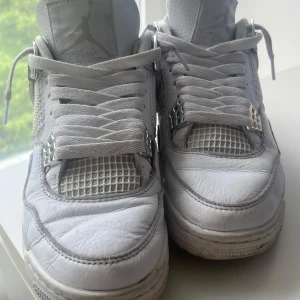 Nike Air Jordans 4 Retro 'Pure Money' vita sneakers - Säljer ett par Nike Air Jordans 4 Retro 'Pure Money' i helvitt läder med silverdetaljer. Klassisk siluett med meshpaneler, snörning och Jumpman-logga på hälen. Perfekta för dig som gillar streetwear och sneakerskultur.