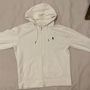 Vit hoodie från Polo Ralph Lauren - Säljer en stilren vit hoodie från Polo Ralph Lauren med dragkedja och huva. Tröjan har fickor framtill och den klassiska broderade loggan på bröstet. Perfekt för en avslappnad look. Lite stor i storleken