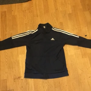 Blå adidas träningsjacka med vita ränder - Mörkblå träningsjacka från adidas med klassiska vita ränder längs ärmarna och dragkedja framtill. Jackan har hög krage och adidas-logga på bröstet. Perfekt för sport eller chill.