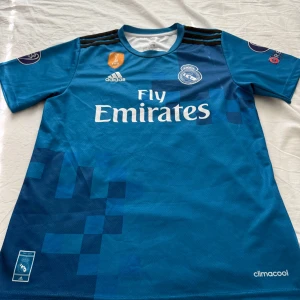 Real Madrid bortatröja Ronaldo 7 - Säljer en blå Real Madrid bortatröja med Ronaldo 7 på ryggen. Tröjan har korta ärmar, vita detaljer och klubbmärke på bröstet. Adidas-logga och Fly Emirates-tryck framtill. Climalite-material för extra komfort.