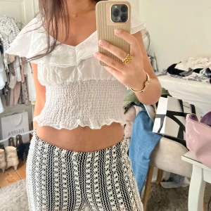 Vit croppad blus med volang - Säljer en söt vit croppad blus med volangdetaljer och smockad midja. Blusen har en v-ringning och är ärmlös, perfekt för varma dagar. Materialet är lätt och luftigt, vilket gör den bekväm att bära. (Obs liten slitning på ena sidan som man ser på sista bilden)