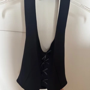 Svart väst från Gina Tricot - Snygg svart väst från Gina Tricot i korsettstil med rosetter framtill. Västen har djup urringning och är figurnära.