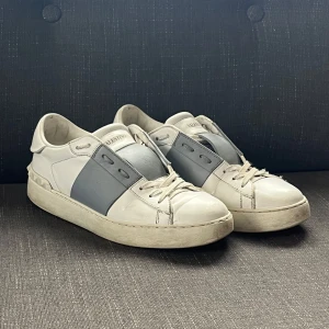 Valentino Open - Säljer ett par Valentino Open sneakers i vitt skinn med grått band över mitten. Klassisk rund tå, platt sula och snörning framtill. Diskret Valentino-logga på plösen och nitar baktill. Perfekta för en stilren och trendig look.