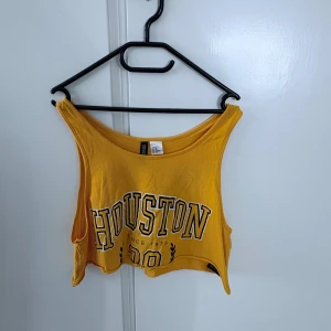 Gul croppad linnetopp med Houston-tryck från Divided - Säljer en gul croppad linnetopp från Divided med stort Houston-tryck framtill. Toppen har breda axelband och är ärmlös, perfekt för varma dagar eller som ett statement-plagg.