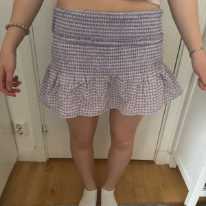 Ljuslila rutig minikjol med volang - Supersöt ljuslila och vit rutig minikjol med volangkant. Kjolen har smock upptill som ger en tight passform och volang nertill för en extra söt look. Perfekt för vår och sommar!