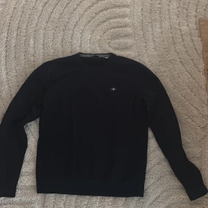 Mörkblå tröja från GANT - Säljer en stilren mörkblå tröja från GANT. Tröjan har rund halsringning, långa ärmar och ribbade muddar vid ärmslut och nederkant. Perfekt för en klassisk och avslappnad look.