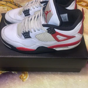 Nike Air Jordan 4 Fire Red - Säljer ett par klassiska Nike Air Jordan 4 Fire Red med vit ovandel, röda och svarta detaljer samt meshpaneler på sidorna. Skorna har den ikoniska Jumpman-loggan på plösen och hälen. Perfekta för dig som gillar streetwear och sneakerskultur.