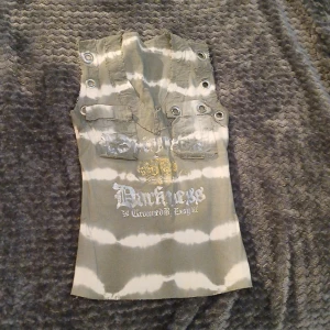 Grå/grön och vit randig ärmlös topp med tryck - Säljer en grå och vit ärmlös topp med knappar och fickdetaljer framtill. Toppen har ett tryck med text och guldfärgad krona på bröstet. Snygg och unik design med ränder och dekorativa knappar vid axlarna.