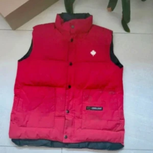 Röd dunväst från Canada Goose - Säljer en röd dunväst från Canada Goose med hög krage och knäppning framtill. Västen har en vit broderad lönnlöv-detalj på bröstet och svart insida. Perfekt för lager-på-lager under kyliga dagar.