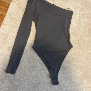 Grå oneshoulder bodysuit - Snygg grå bodysuit med oneshoulder-design och lång ärm. Perfekt för att skapa en trendig och stilren look. Tight passform som framhäver figuren.