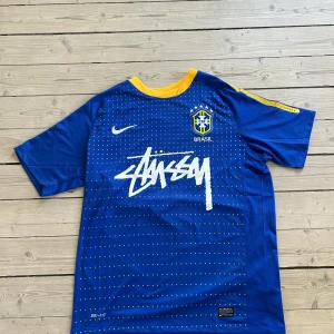Blå Brasil fotbollströja Nike x Stussy - Säljer en blå Brasil fotbollströja från Nike i samarbete med Stussy. Tröjan har korta ärmar, gul krage och detaljer, samt vit Stussy-logga på bröstet. Materialet är Dri-Fit för bra ventilation. Perfekt för fotboll eller streetwear. Brasilien