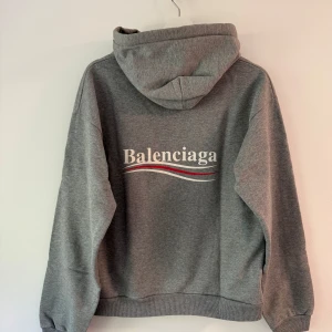 Grå hoodie från Balenciaga (med kvitto) - Säljer en grå hoodie från Balenciaga med logga tryckt på bröstet och ryggen. Tröjan har huva med dragsko och en stor magficka. Lite kortare i kroppen med en lätt boxigare passform.