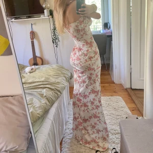 Vit långklänning med blommor och knytband - Supersöt långklänning i vit med rosa blommigt mönster. Klänningen har smala axelband, knytning framtill och jättesnygg figurnära passform. Perfekt för sommaren då materialet känns mjukt och lätt💞 Den har lite ”sjöjungfru” modell då den är lite vidare där nere😍 