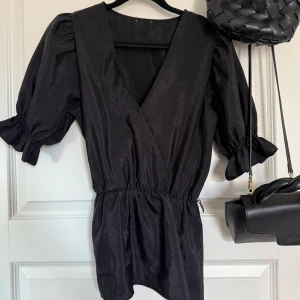 Svart omlottblus med puffärm - Snygg svart omlottblus med djup v-ringning och puffiga kortärmar. Blusen har resår i midjan för en smickrande passform och är tillverkad i ett glansigt material. Perfekt för dig som gillar stilrena och trendiga plagg. 