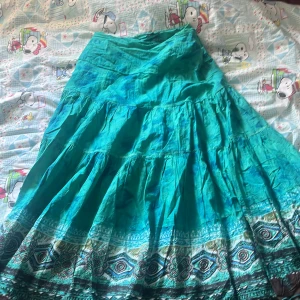 Turkos långkjol med mönster från effgee - Säljer en turkos långkjol från effgee med vackert etno-inspirerat mönster i blått, svart och vitt längs nederkanten. Kjolen har en luftig och svepande passform, dragkedja i sidan och är tillverkad i bomull.