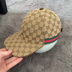 Beige keps från Gucci med GG-mönster - Snygg beige keps från Gucci med klassiskt GG-mönster i brunt och grönt/rött band på sidan. Justerbar rem baktill i brunt skinn. Perfekt accessoar för en trendig look. + Gucci påse