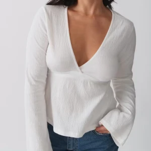 Vit omlottopp med v-ringning - Säljer en vit omlottopp med djup v-ringning och långa ärmar. Toppen har en knytning i midjan och är tillverkad i ett lätt, strukturerat material. Perfekt att styla med jeans eller kjol.  Väldigt bra skick!