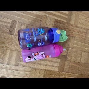 Två vattenflaskor för barn med djurmotiv - Säljer två färgglada vattenflaskor från Nalgene för barn. En är lila/blå med grönt lock och robotmotiv, den andra är rosa med djurmotiv och rosa lock. Båda har praktiska piplock och är tillverkade i tålig plast.