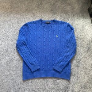 Blå kabelstickad tröja från Polo Ralph Lauren - Pris kan diskuteras! Säljer en blå kabelstickad tröja från Polo Ralph Lauren med rund halsringninglogga. Tröjan har långa ärmar och ribbade muddar vid ärmslut och nederkant. Perfekt för lager-på-lager-stil.