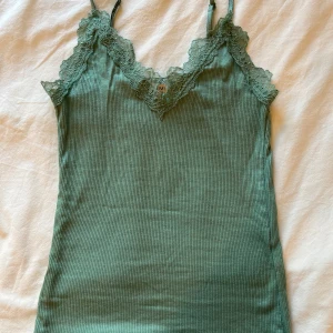 Grönt linne från New Yorker  - Säljer ett grönt linne fårn New Yorker i storlek XXS.  Linnet har fina spetsdetaljer längs urringningen och axelbanden. Perfekt att styla till jeans eller kjol för en fräsch look.