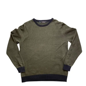 Dressmann tröja - Säljer en olivgrön stickad tröja från Dressmann med mörkgrå ribbade muddar vid hals, ärmslut och nederkant. Tröjan är tillverkad i bomull och har en klassisk rund halsringning. Perfekt för lager-på-lager under kyligare dagar. Tröjan har inga tecken på användning.