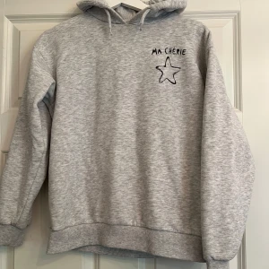 Grå hoodie med tryck 'Ma Chérie' - Säljer en ljusgrå hoodie med huva och trycket 'Ma Chérie' samt en stjärna både fram och bak. Tröjan har långa ärmar och ribbade muddar vid ärmslut och nederkant. Perfekt för en avslappnad stil. Nypris: 250kr