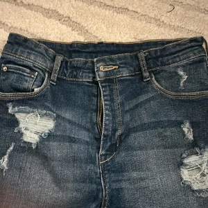 Mörkblå slitna jeans shorts  - Säljer ett par mörkblå jeans shorts med slitna detaljer och dragkedja fram. Shortsen har klassisk femficksdesign och bälteshällor. Perfekta för en avslappnad och trendig look.