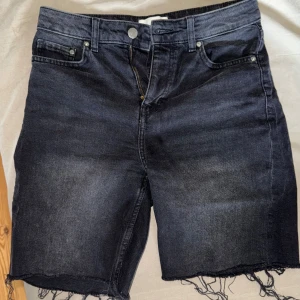 Svarta jeansshorts med fransig kant - Snygga svarta jeansshorts med hög midja och rå, fransig kant nertill. Klassisk shorts med knapp och dragkedja framtill. Perfekta för en avslappnad stil. Perfekta för sommaren. 
