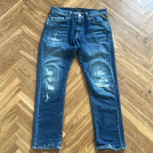Replay Mini Waitom jeans med slitningar - Mörkblå jeans från Replay, modell Mini Waitom. Klassisk femficksdesign med raka ben och slitna detaljer på både fram- och baksida. Jeansen har knappgylf och tydliga Replay-loggor på fickorna. Perfekta för en avslappnad stil.