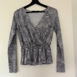 Svartvit omlottopp med paisleymönster från Bik Bok - Säljer en svartvit omlottopp från Bik Bok med paisleyinspirerat mönster. Toppen har lång ärm, v-ringning och markerad midja med lätt volang nedtill. Knappt använd 